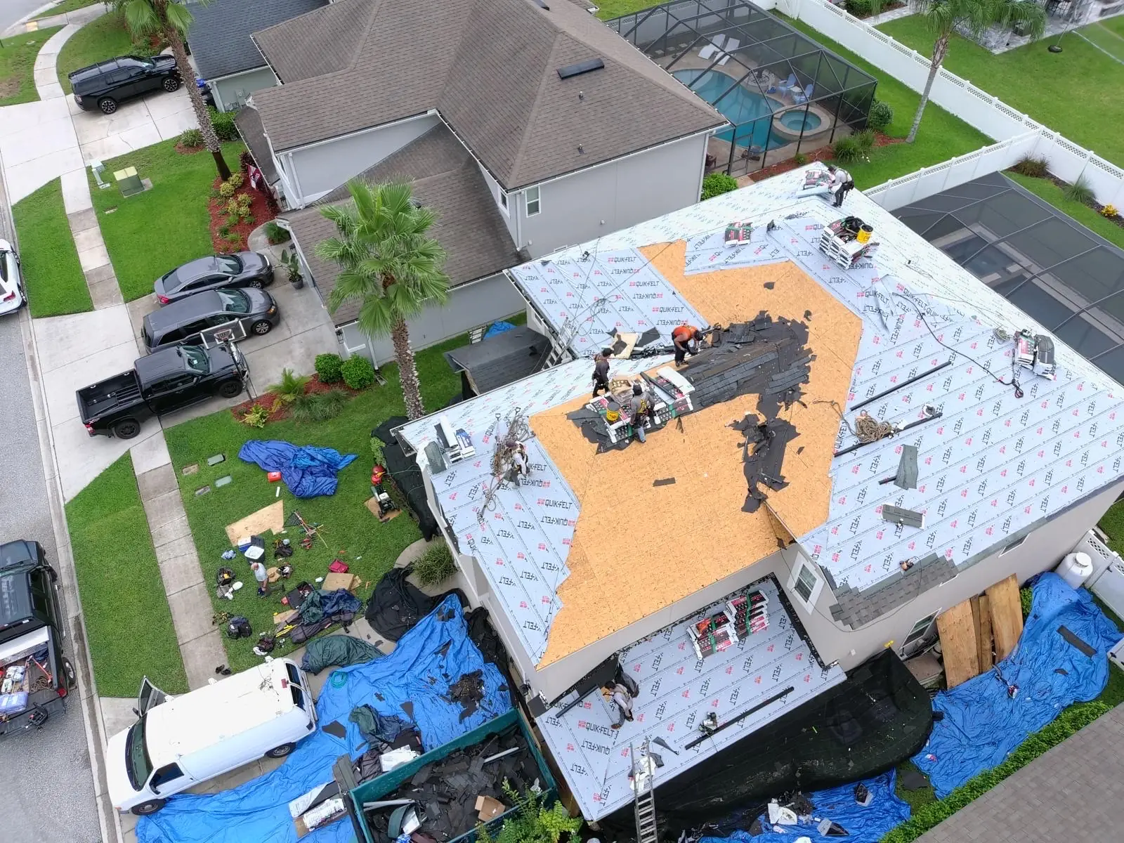 Aerial view of a roof tear-off in progress beside a screened pool, with plywood decking exposed
