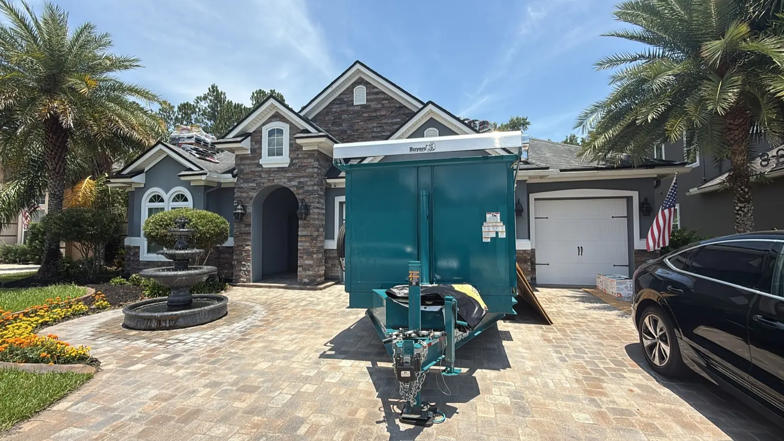Stone-front home with a fountain and palm trees, Miller Roofing dump trailer staged in the driveway