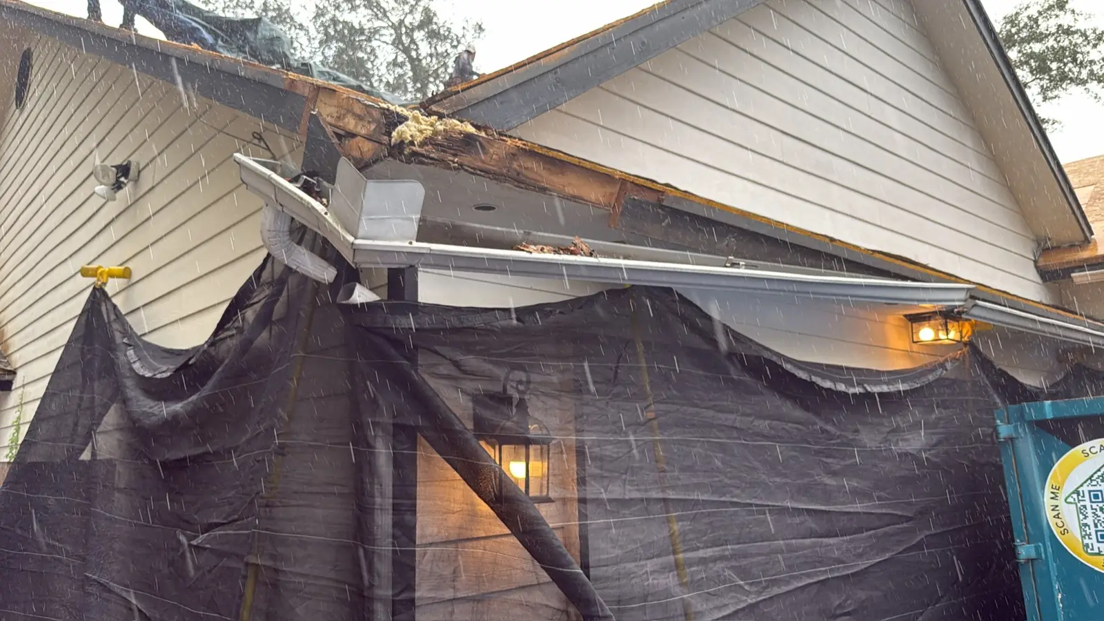 Rain falling on a home with storm-damaged fascia and a hanging gutter, protective screens over the entry