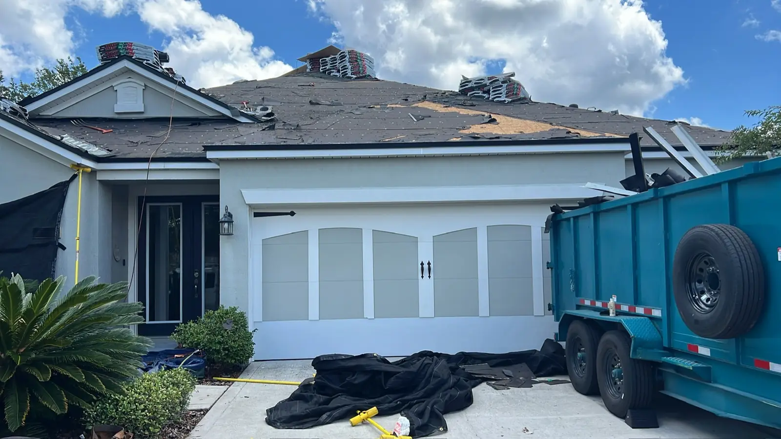 Front of a home mid tear-off with bundles staged on the roof and a dump trailer at the garage