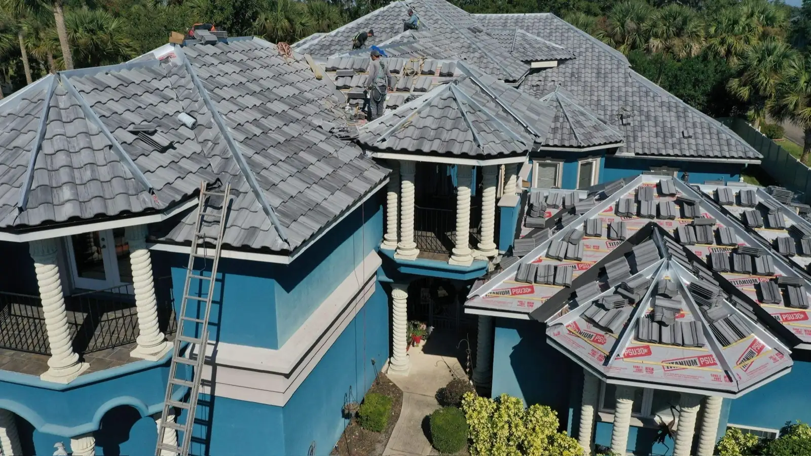 Drone view of a concrete tile re-roof in progress on a large two-story home with columns and palm trees